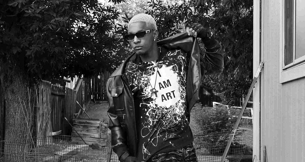 Person with sunglasses and blonde hair, wearing jacket over "I AM ART" shirt, stands confidently outdoors with trees and a fence in the background.