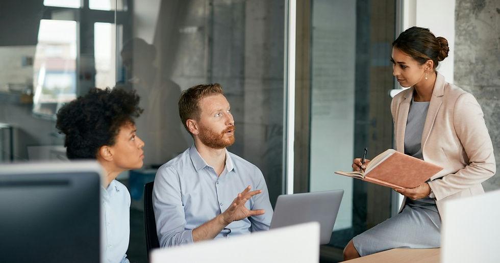 Three coworkers in a glass office meeting, one woman taking notes while a man speaks and another woman listens attentively