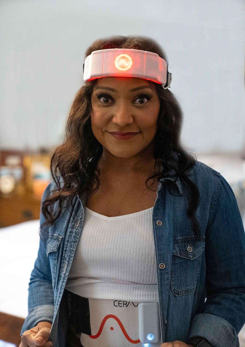 Woman in a denim jacket smiles, wearing a glowing red headband and a device labeled "Cera", in a cozy indoor setting.