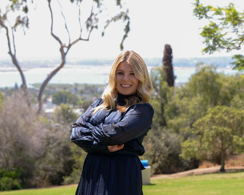 Smiling woman in a dark dress, arms crossed, standing in a park with trees and distant water view, under a bright, sunny sky.