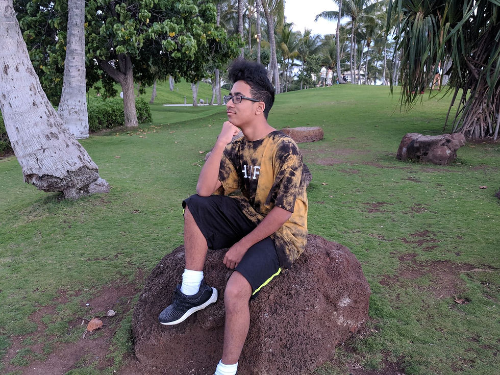 Young man in glasses sits thoughtfully on a rock in a tropical park, wearing a black-and-gold tie-dye shirt and shorts.