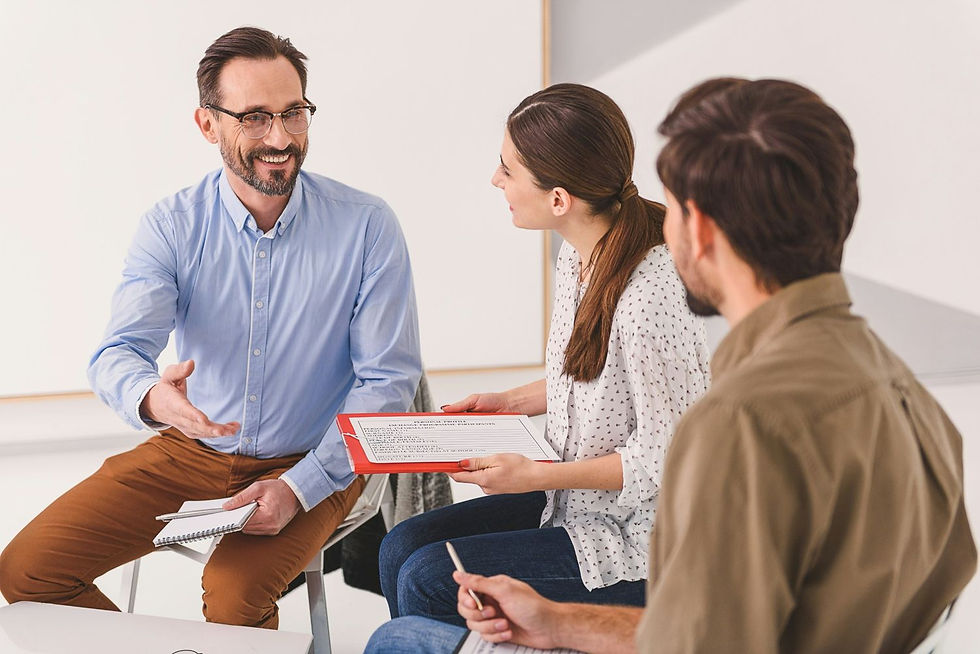 Three people are in discussion in a bright room. A man extends his hand, smiling. Another holds a clipboard with visible text. Casual setting.