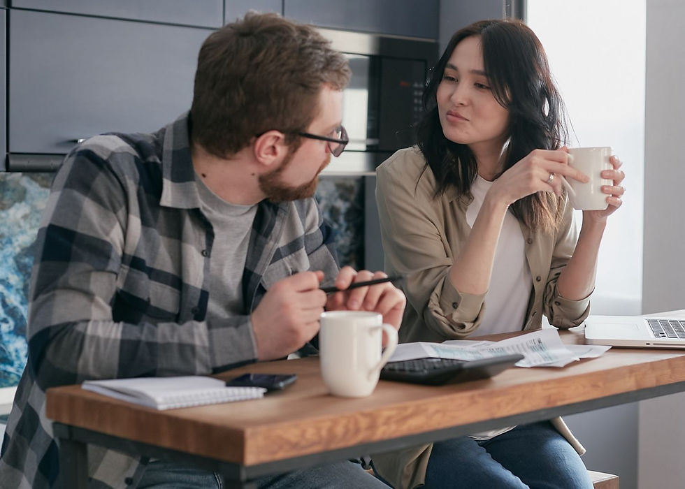 Two people talk at a kitchen table with mugs, papers, and a laptop, looking serious and focused.