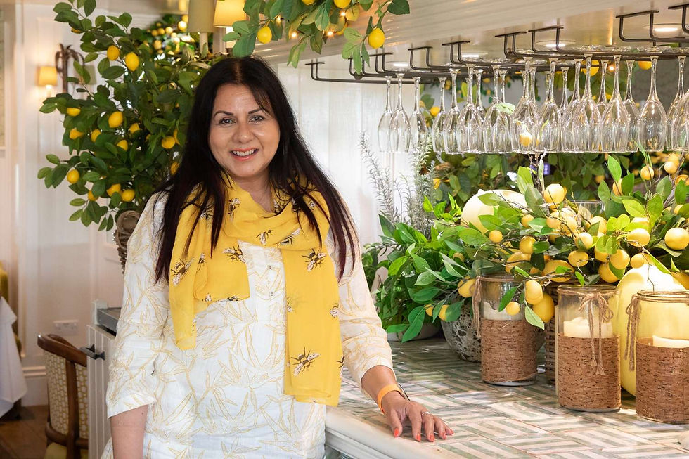 Smiling woman in a lemon-decorated bar, with hanging wine glasses, candles, and lush greenery in a bright indoor setting.