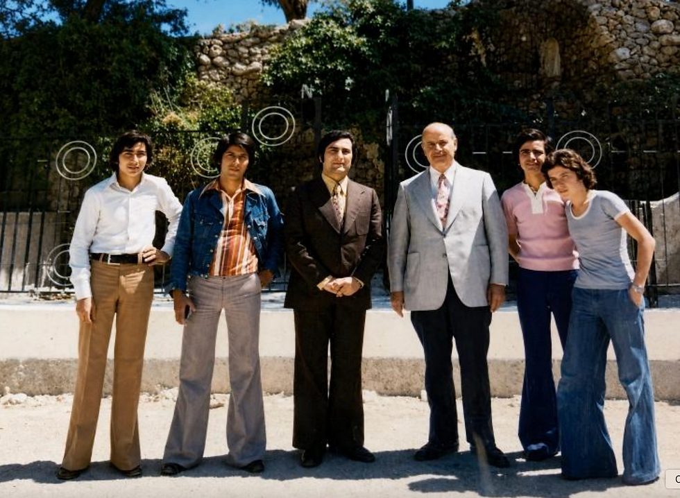 Six men pose outdoors in front of a stone wall and iron gate, wearing 1970s-style clothes in a sunny group portrait.
