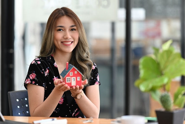 Smiling woman in a floral dress holds a small red house model at a desk in a bright office setting.