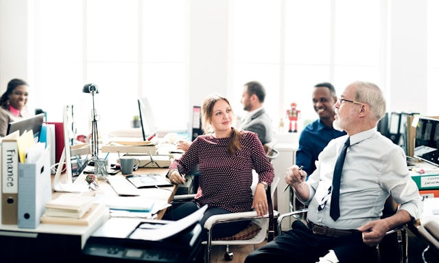 Office setting with five people conversing and smiling at desks. Bright windows in the background. A binder labeled "Finance" on the desk.