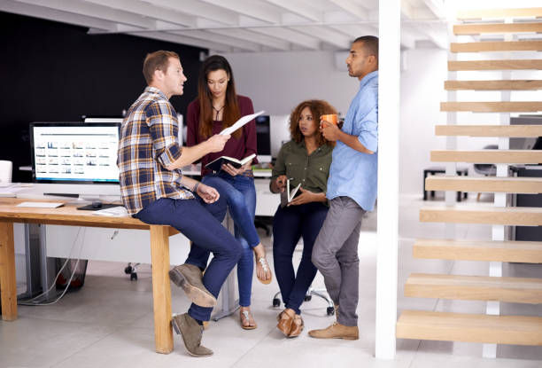 Four people in casual attire have a meeting in a modern office near a staircase. One holds papers; another has a mug. A computer is visible.