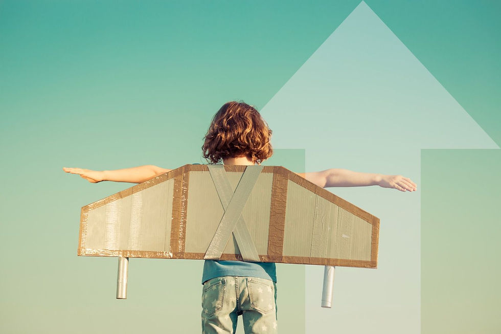 Child with cardboard wings stands, arms outstretched against a blue sky, suggesting flight. Wearing star-patterned pants, evoking dreams.