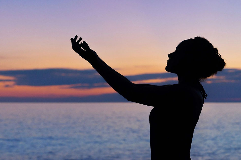 Silhouette of a person with raised hands against a colorful sunset sky by the sea, creating a peaceful and contemplative mood.