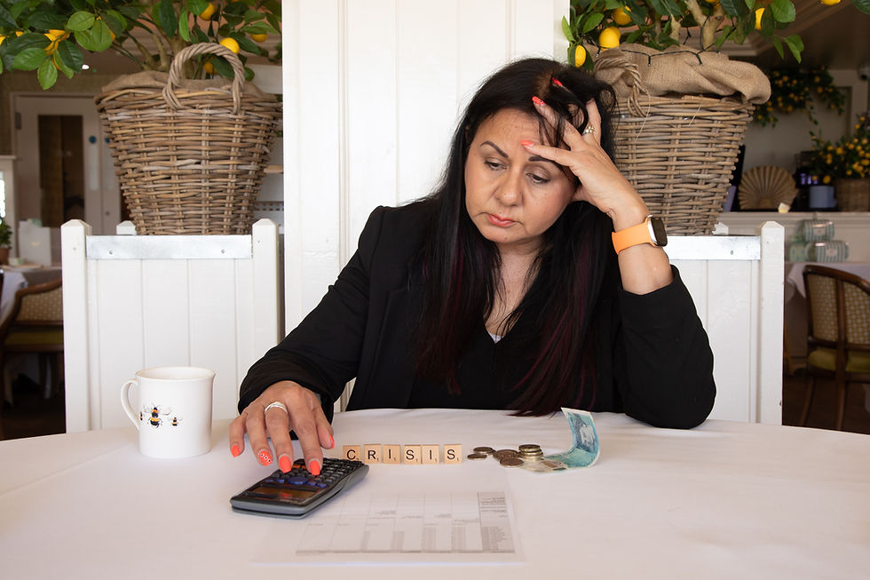Woman looks stressed at a table, pressing a calculator beside crisis tiles, coins, and a mug in a bright cafe.