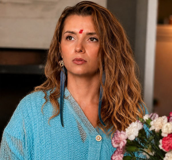 Woman in a blue top with a red mark on her forehead looks worried indoors, with flowers in the foreground.