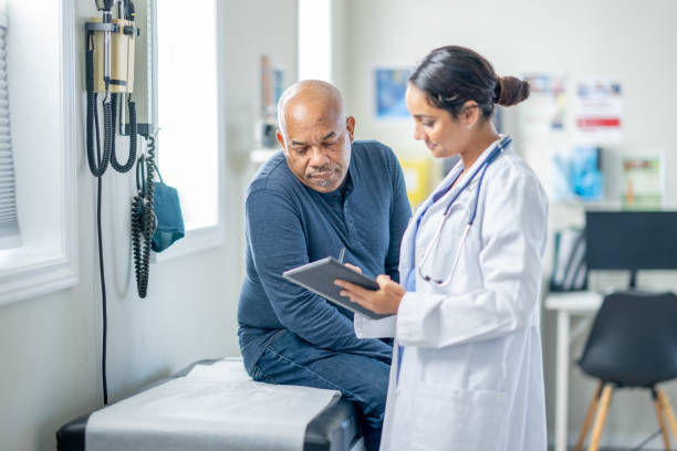 A senior gentleman sits up on an exam table during a routine check-up. His female doctor is holding out a tablet as they review some recent test results together.