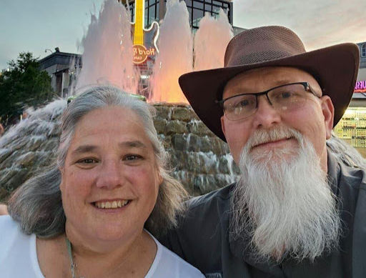 Smiling couple in front of a lit fountain, one wearing glasses and a hat. Neon guitar sign in background suggests evening setting.