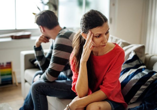 Tense couple sits apart on a couch in a living room; woman in red rubs her temple while man looks away.