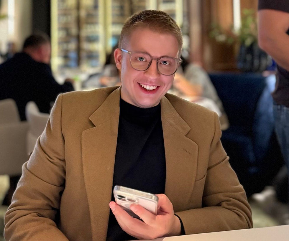 Man in a tan jacket smiling and holding a phone in a dimly lit restaurant. Blurred background with people and soft lighting.