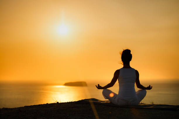 Silhouette of a person meditating at sunset on a cliff overlooking the sea. The sky and ocean glow with warm orange hues, creating a serene mood.