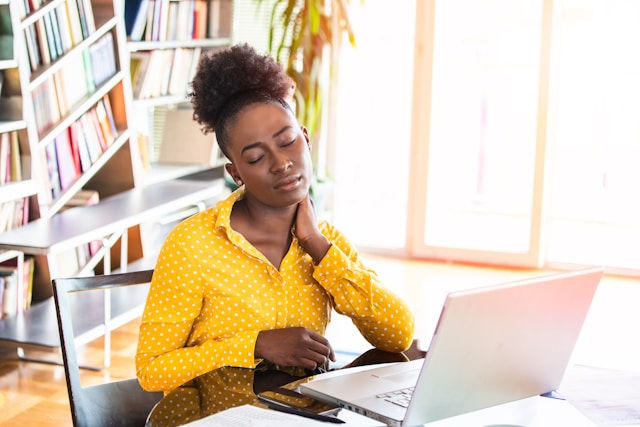 Woman in a yellow polka dot shirt sits at a desk with a laptop, hand on neck, eyes closed, appearing tired. Bookshelves in the background.