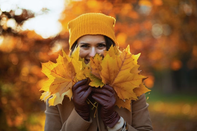 Woman in a yellow beanie smiles behind a bouquet of golden autumn leaves, with a warm orange fall background.