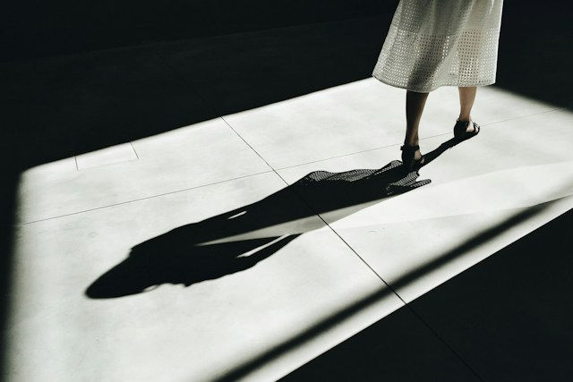 Lower half of a woman in a white dress stands on a bright floor, casting a long dark shadow in a moody, minimal space.