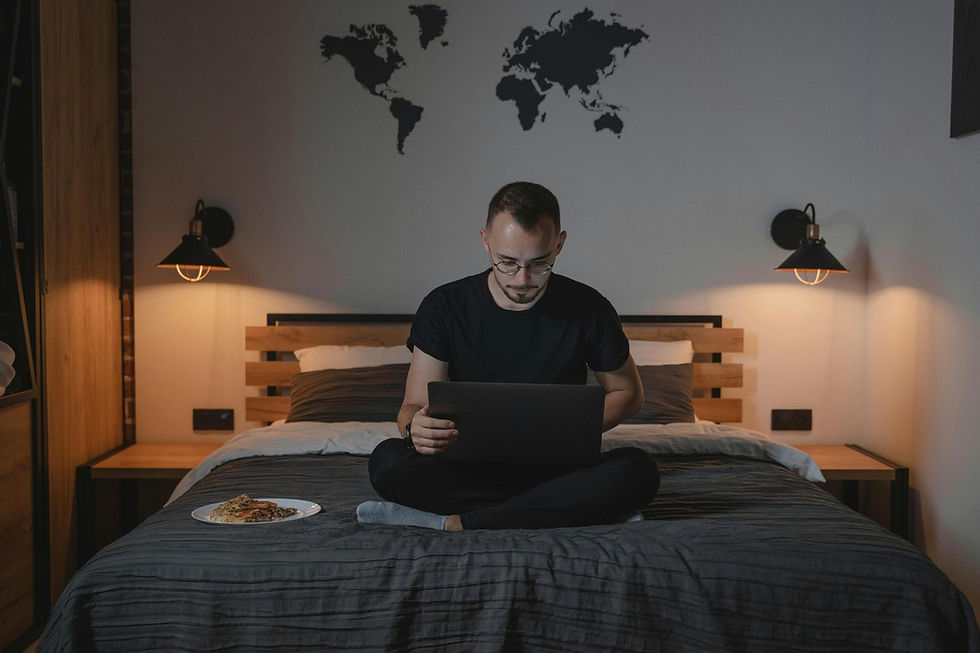 Man sitting on bed with laptop, focused. Dim room with world map on wall, warm lighting, and a plate of food beside him. Cozy mood.