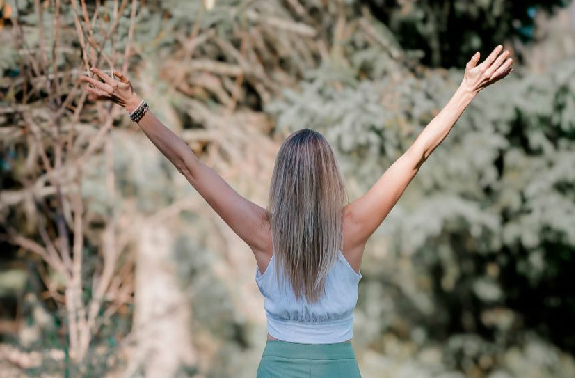 Woman with arms raised in a forest setting, wearing a white top and green pants, exuding a sense of freedom and positivity.