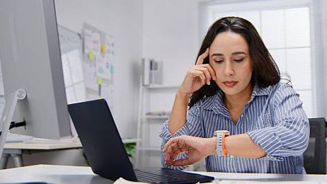 Woman in a blue striped shirt checks her watch beside a laptop in a bright office, looking focused and slightly impatient.