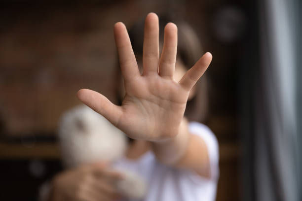Child holds out hand to camera, obscuring face. Holds plush toy in other hand. Blurry indoor background, suggesting a rejecting gesture.