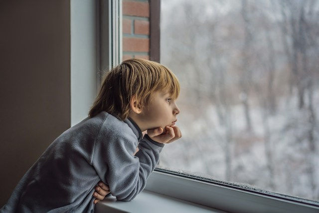 Young child in a gray sweater leans on a windowsill, gazing out at a snowy blurred landscape with a quiet, wistful mood.