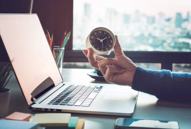 Hand holding a small alarm clock beside an open laptop on a desk, with a bright window and city blur behind