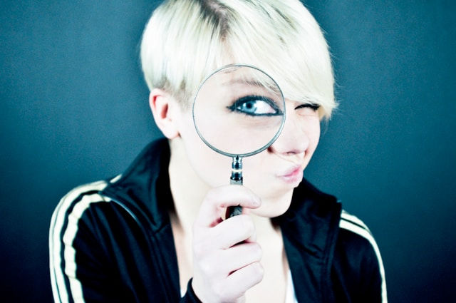 A woman with short blonde hair holding a magnifying glass over one eye, making it appear enlarged.