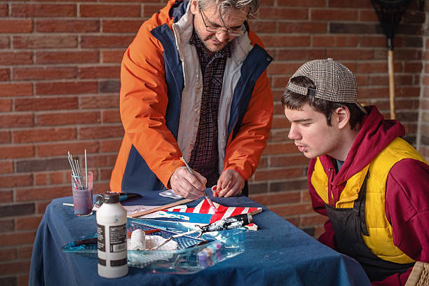 Older man paints a red, white and blue design on a table while a young man watches, both focused beside a brick wall.