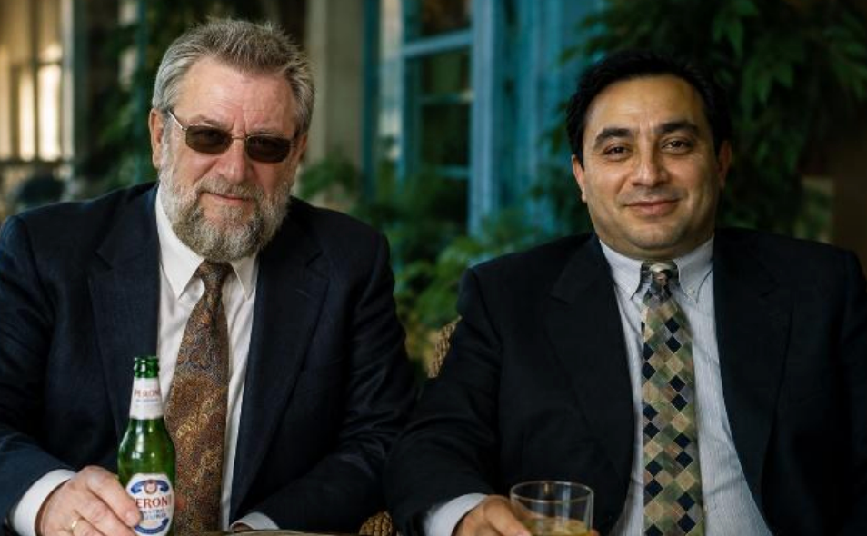 Two suited men sit smiling indoors; one wears sunglasses and holds a Peroni beer, the other a drink, with greenery behind.