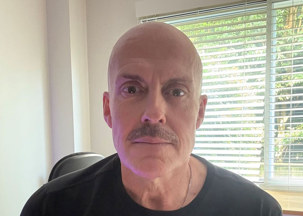 Bald man with a mustache looks at the camera indoors, in front of bright window blinds, serious expression.