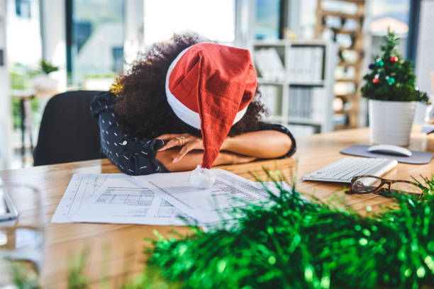 Person in Santa hat asleep on office desk with papers, surrounded by holiday decor. Bright daylight and festive mood.