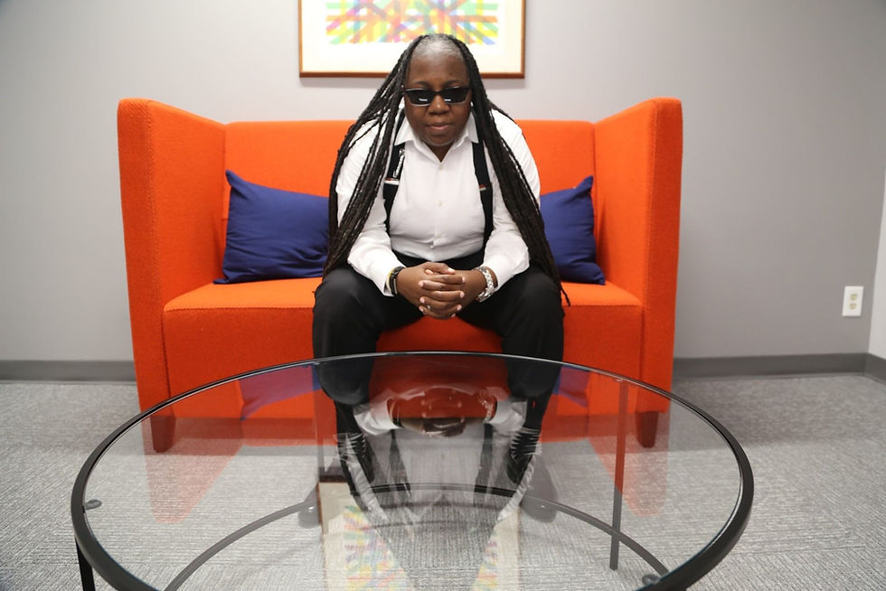 Person in sunglasses sits on an orange couch with blue pillows, leaning forward in a modern office lounge behind a glass table.
