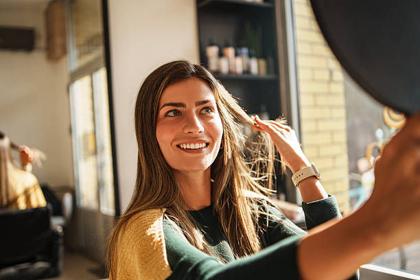 Smiling woman in a salon chair as a stylist adjusts her hair in warm sunlight, with shelves and a brick wall behind her.