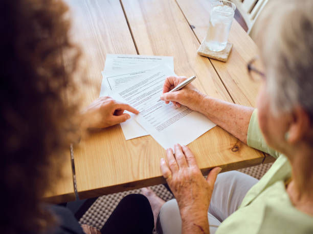 Elderly woman signs a document at a wooden table while another person points; glass of water nearby.