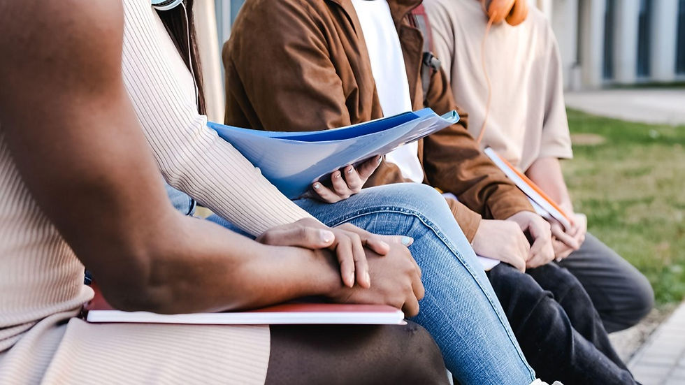 Students sit outdoors on campus with notebooks in their laps, hands clasped, appearing to study or listen together.