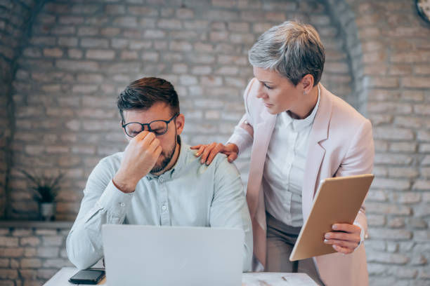 Concerned woman comforts stressed man at a laptop in a brick-walled office, her hand on his shoulder, holding a tablet.
