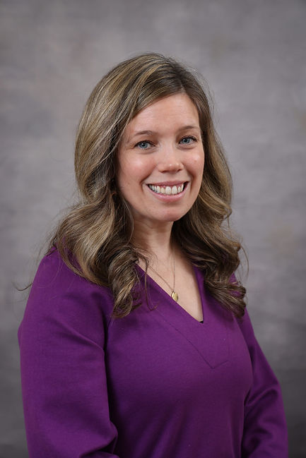 A woman with long hair smiles in a purple top. She stands against a gray background, exuding a friendly and approachable vibe.