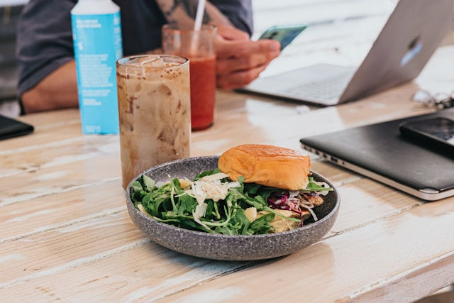 Salad and sandwich on a wooden table with iced drinks; person using a smartphone near a laptop in a casual, bright setting.