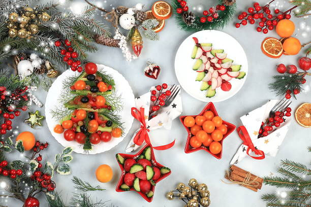 Festive table with fruit and veggie holiday trees, red and orange decor, surrounded by pine branches and berries. Bright, joyful mood.