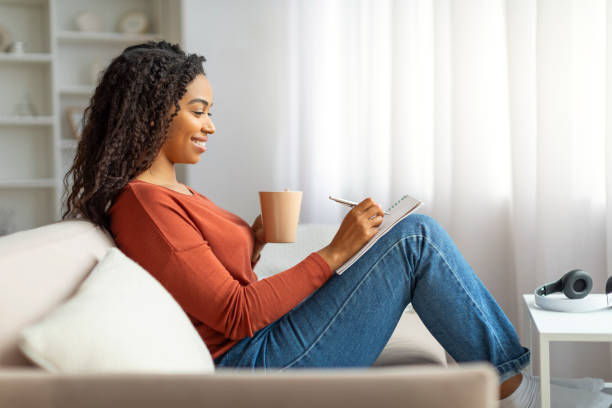 A woman sits comfortably on a sofa, smiling as she journals in a notebook while holding a mug, with natural light streaming through a nearby window.