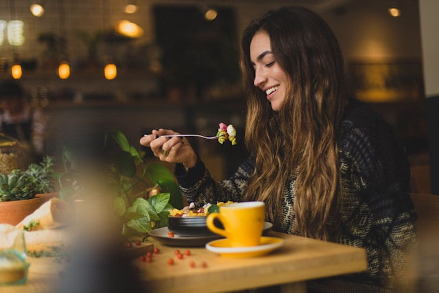 Smiling woman enjoys a meal at a cozy café, with warm lighting. Yellow cup and greenery on the table; relaxed and cheerful atmosphere.