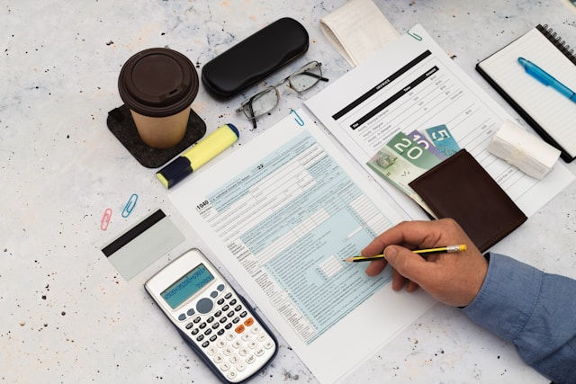 A person is reviewing financial documents at a desk, surrounded by a calculator, coffee cup, money, credit card, glasses, and stationery.