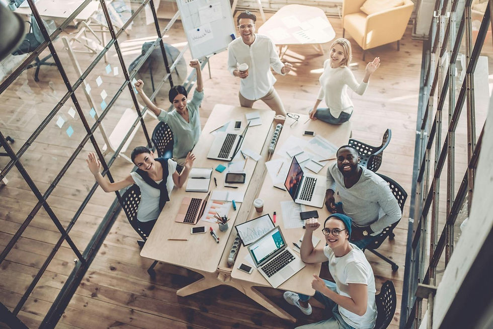 Aerial view of six people in a bright office cheering around a table with laptops. Wooden floor, glass walls, and a chart board in background.