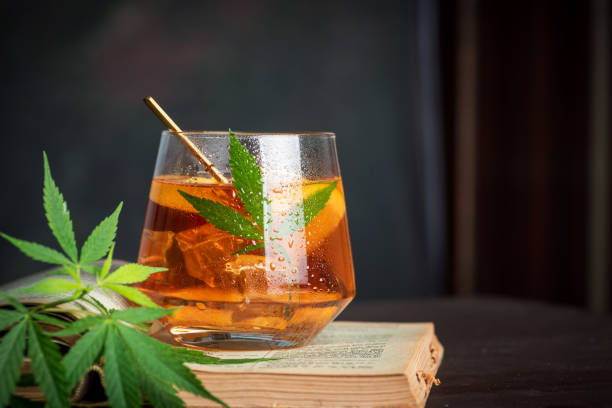 A glass of iced tea with a cannabis leaf garnishing it sits on an open book, against a dark background. Mood is relaxed, colors are warm.