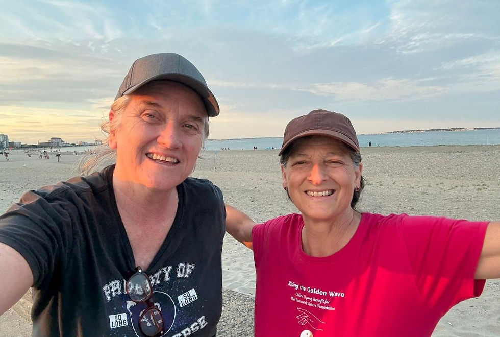 Two people smiling on a beach at sunset. One wears a black shirt with text, the other a red shirt and brown cap. Ocean and sky in background.