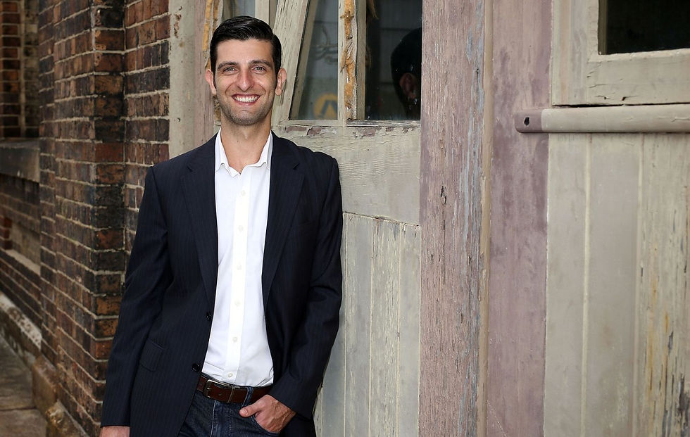 Smiling man in a dark blazer and white shirt leans by a weathered brick and wood wall in a rustic alley.